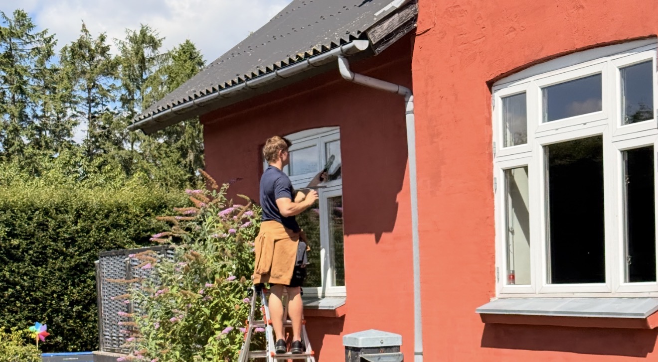 Marius pudser et vindue med skvis på en stige ved et rødt hus på Fyn